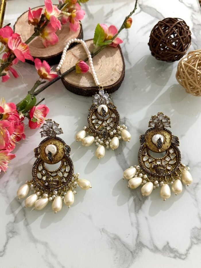 Antique gold maang tikka and earring set with kundan stones and pearl drop embellishments, displayed on a marble surface with floral and wooden decor accents.