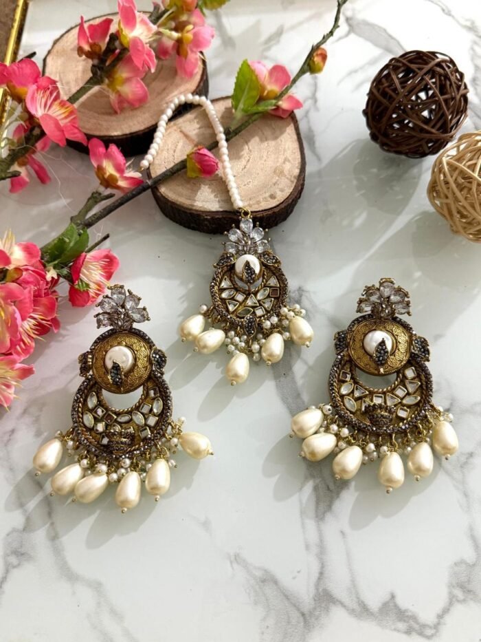 Antique gold maang tikka and earring set with kundan stones and pearl drop embellishments, displayed on a marble surface with floral and wooden decor accents.