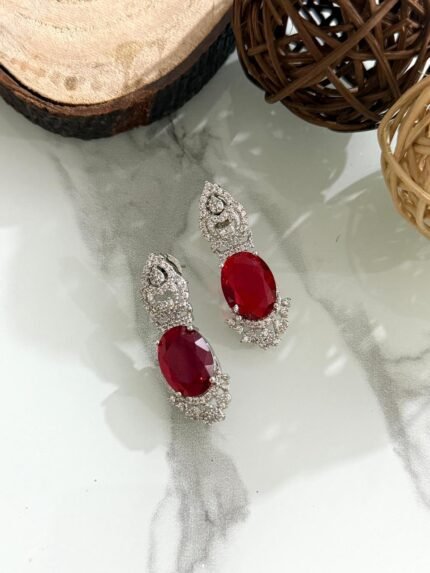 Three pairs of drop earrings with red, green, and blue oval stones, accented by sparkling diamond-cut silver designs, placed on a white marble surface.