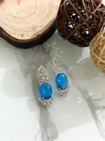 Three pairs of drop earrings with red, green, and blue oval stones, accented by sparkling diamond-cut silver designs, placed on a white marble surface.