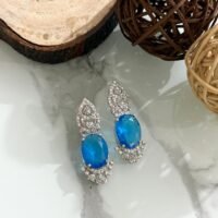 Three pairs of drop earrings with red, green, and blue oval stones, accented by sparkling diamond-cut silver designs, placed on a white marble surface.