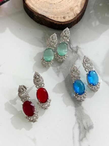 Three pairs of drop earrings with red, green, and blue oval stones, accented by sparkling diamond-cut silver designs, placed on a white marble surface.