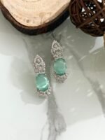 Three pairs of drop earrings with red, green, and blue oval stones, accented by sparkling diamond-cut silver designs, placed on a white marble surface.