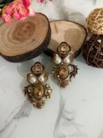 Two pairs of ornate traditional earrings with red and amber center stones, teardrop pearls, and gold detailing placed on a marble surface with decorative wooden props.
