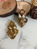 Two pairs of ornate traditional earrings with red and amber center stones, teardrop pearls, and gold detailing placed on a marble surface with decorative wooden props.