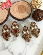 Two pairs of ornate traditional earrings with red and amber center stones, teardrop pearls, and gold detailing placed on a marble surface with decorative wooden props.