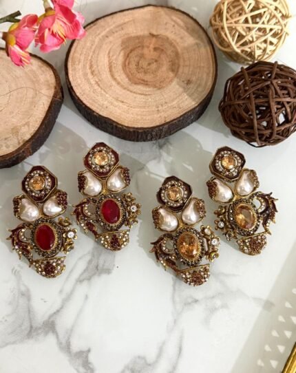 Two pairs of ornate traditional earrings with red and amber center stones, teardrop pearls, and gold detailing placed on a marble surface with decorative wooden props.