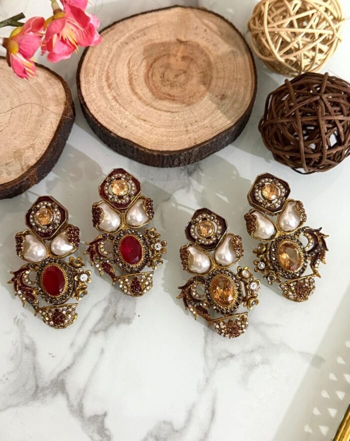 Two pairs of ornate traditional earrings with red and amber center stones, teardrop pearls, and gold detailing placed on a marble surface with decorative wooden props.