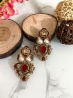 Two pairs of ornate traditional earrings with red and amber center stones, teardrop pearls, and gold detailing placed on a marble surface with decorative wooden props.