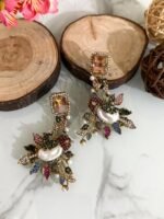 A set of ornate, handcrafted earrings displayed on a marble surface with decorative wood slices and floral accents. Each earring features a large gem stud, pearl centerpiece, and intricate leaf motifs adorned with multicolored stones.