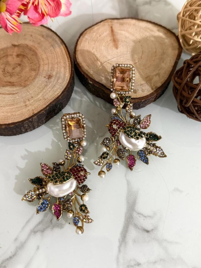 A set of ornate, handcrafted earrings displayed on a marble surface with decorative wood slices and floral accents. Each earring features a large gem stud, pearl centerpiece, and intricate leaf motifs adorned with multicolored stones.
