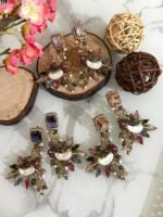 A set of ornate, handcrafted earrings displayed on a marble surface with decorative wood slices and floral accents. Each earring features a large gem stud, pearl centerpiece, and intricate leaf motifs adorned with multicolored stones.