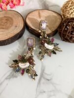 A set of ornate, handcrafted earrings displayed on a marble surface with decorative wood slices and floral accents. Each earring features a large gem stud, pearl centerpiece, and intricate leaf motifs adorned with multicolored stones.