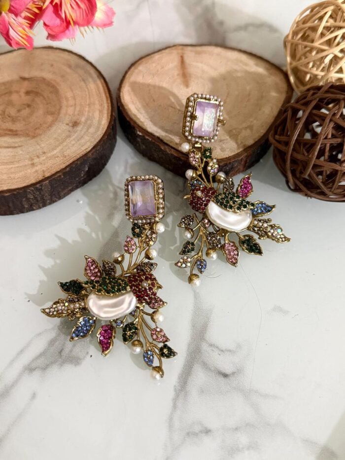 A set of ornate, handcrafted earrings displayed on a marble surface with decorative wood slices and floral accents. Each earring features a large gem stud, pearl centerpiece, and intricate leaf motifs adorned with multicolored stones.