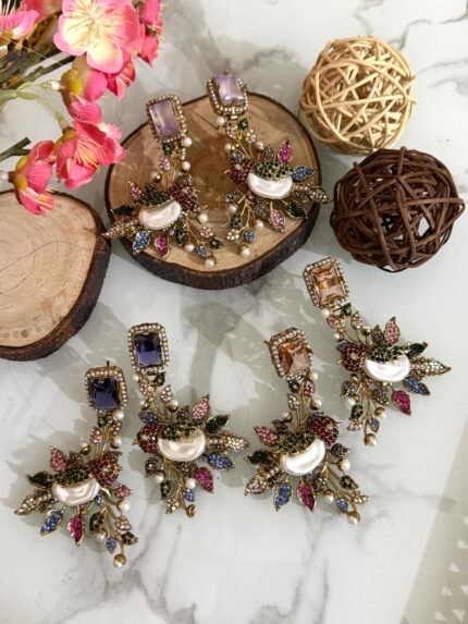 A set of ornate, handcrafted earrings displayed on a marble surface with decorative wood slices and floral accents. Each earring features a large gem stud, pearl centerpiece, and intricate leaf motifs adorned with multicolored stones.