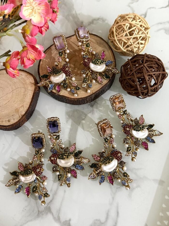A set of ornate, handcrafted earrings displayed on a marble surface with decorative wood slices and floral accents. Each earring features a large gem stud, pearl centerpiece, and intricate leaf motifs adorned with multicolored stones.