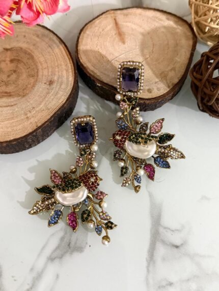 A set of ornate, handcrafted earrings displayed on a marble surface with decorative wood slices and floral accents. Each earring features a large gem stud, pearl centerpiece, and intricate leaf motifs adorned with multicolored stones.