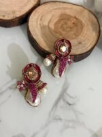A stylish assortment of embellished pearl drop stud earrings in various jewel tones including red, green, purple, and gold, displayed on a marble surface with rustic wood slices and decorative wicker balls.