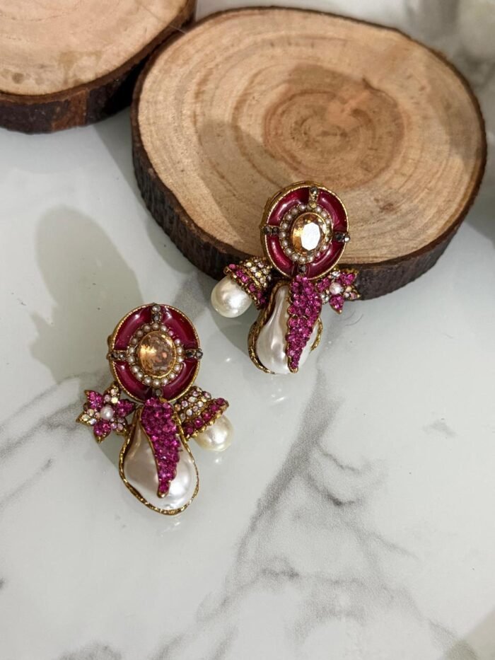 A stylish assortment of embellished pearl drop stud earrings in various jewel tones including red, green, purple, and gold, displayed on a marble surface with rustic wood slices and decorative wicker balls.