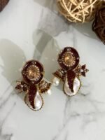 A stylish assortment of embellished pearl drop stud earrings in various jewel tones including red, green, purple, and gold, displayed on a marble surface with rustic wood slices and decorative wicker balls.
