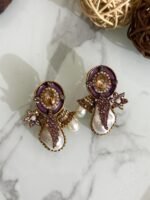 A stylish assortment of embellished pearl drop stud earrings in various jewel tones including red, green, purple, and gold, displayed on a marble surface with rustic wood slices and decorative wicker balls.