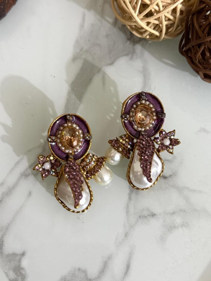 A stylish assortment of embellished pearl drop stud earrings in various jewel tones including red, green, purple, and gold, displayed on a marble surface with rustic wood slices and decorative wicker balls.