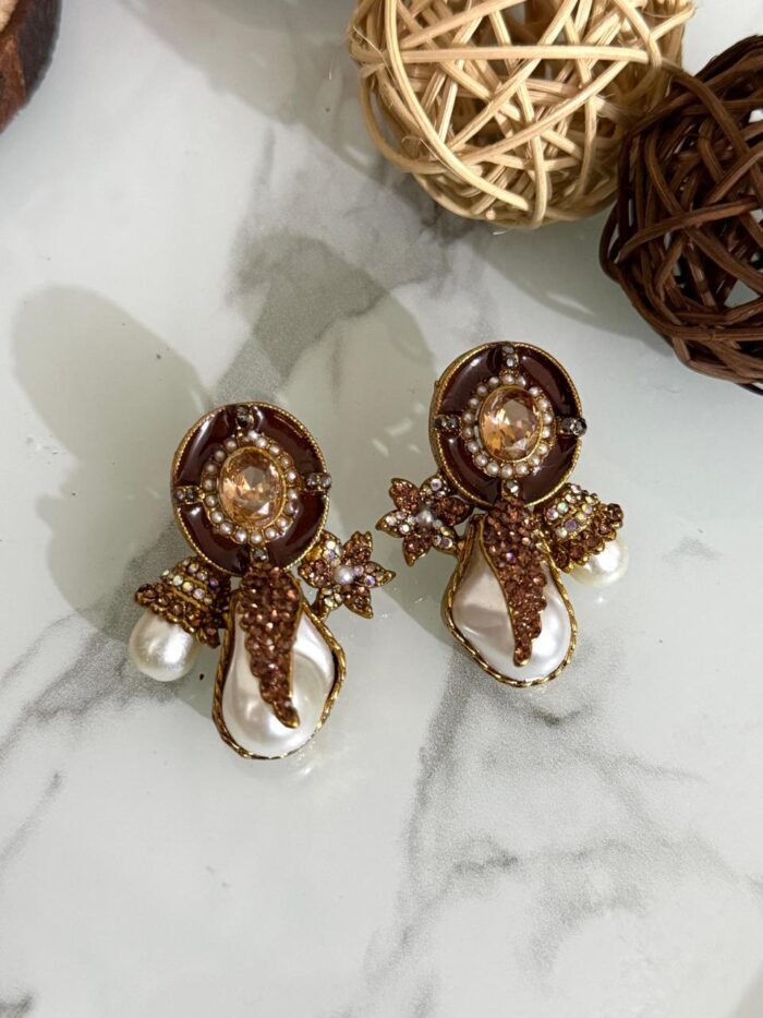 A stylish assortment of embellished pearl drop stud earrings in various jewel tones including red, green, purple, and gold, displayed on a marble surface with rustic wood slices and decorative wicker balls.