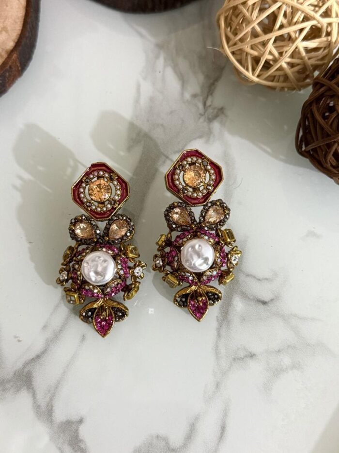 A luxurious collection of ornate antique-finish earrings, each featuring layered floral designs with crystals, faux pearls, and jewel-toned accents, displayed on a marble surface with wood and wicker decor