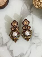 A luxurious collection of ornate antique-finish earrings, each featuring layered floral designs with crystals, faux pearls, and jewel-toned accents, displayed on a marble surface with wood and wicker decor