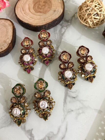 A luxurious collection of ornate antique-finish earrings, each featuring layered floral designs with crystals, faux pearls, and jewel-toned accents, displayed on a marble surface with wood and wicker decor