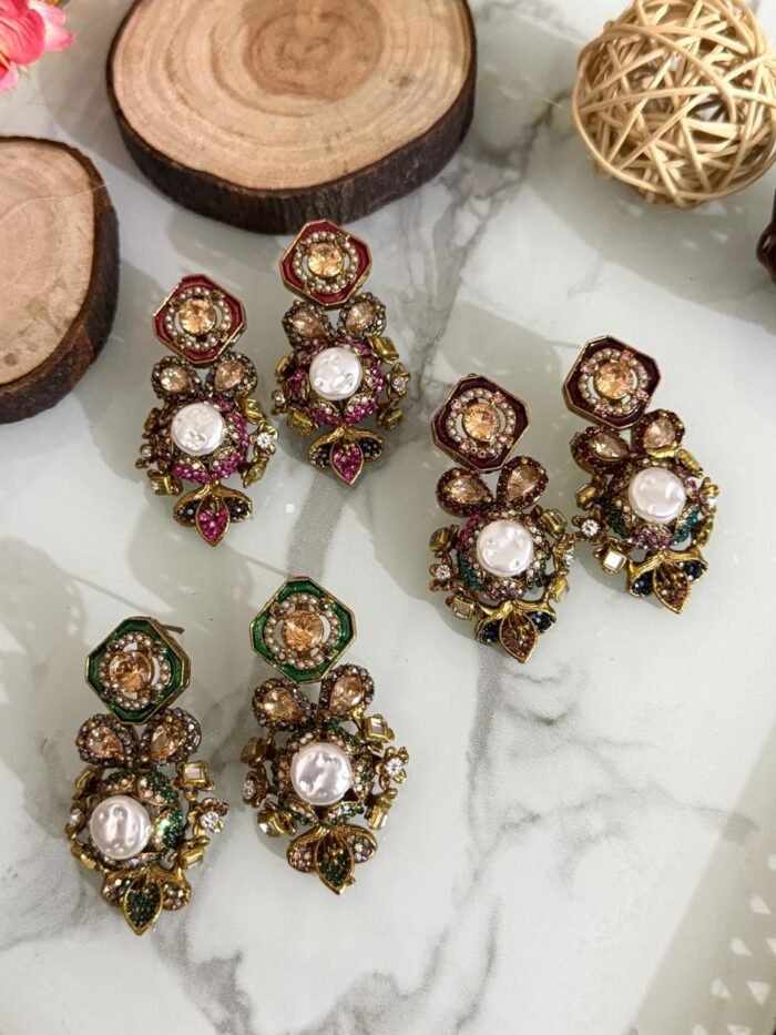 A luxurious collection of ornate antique-finish earrings, each featuring layered floral designs with crystals, faux pearls, and jewel-toned accents, displayed on a marble surface with wood and wicker decor