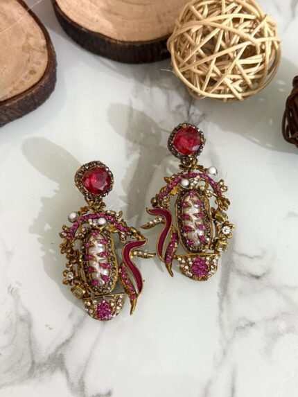 A vibrant collection of statement drop earrings with pearl-embroidered centers, ornate golden frames, and jewel-toned accents in black, teal, amber, red, and pink — displayed on a marble surface with wooden and wicker decor.