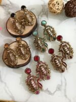 A vibrant collection of statement drop earrings with pearl-embroidered centers, ornate golden frames, and jewel-toned accents in black, teal, amber, red, and pink — displayed on a marble surface with wooden and wicker decor.