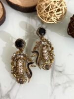 A vibrant collection of statement drop earrings with pearl-embroidered centers, ornate golden frames, and jewel-toned accents in black, teal, amber, red, and pink — displayed on a marble surface with wooden and wicker decor.