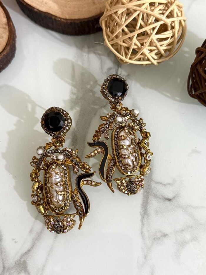 A vibrant collection of statement drop earrings with pearl-embroidered centers, ornate golden frames, and jewel-toned accents in black, teal, amber, red, and pink — displayed on a marble surface with wooden and wicker decor.