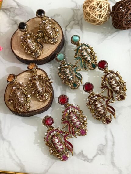 A vibrant collection of statement drop earrings with pearl-embroidered centers, ornate golden frames, and jewel-toned accents in black, teal, amber, red, and pink — displayed on a marble surface with wooden and wicker decor.