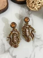 A vibrant collection of statement drop earrings with pearl-embroidered centers, ornate golden frames, and jewel-toned accents in black, teal, amber, red, and pink — displayed on a marble surface with wooden and wicker decor.