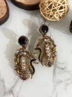 A vibrant collection of statement drop earrings with pearl-embroidered centers, ornate golden frames, and jewel-toned accents in black, teal, amber, red, and pink — displayed on a marble surface with wooden and wicker decor.