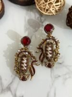A vibrant collection of statement drop earrings with pearl-embroidered centers, ornate golden frames, and jewel-toned accents in black, teal, amber, red, and pink — displayed on a marble surface with wooden and wicker decor.