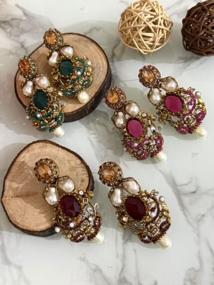 A rich collection of ornate drop earrings featuring oversized gemstones, baroque-style pearls, and detailed gold filigree with colored stone accents — arranged on a marble surface with natural wood and wicker decor.