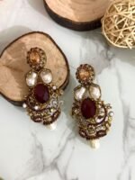 A rich collection of ornate drop earrings featuring oversized gemstones, baroque-style pearls, and detailed gold filigree with colored stone accents — arranged on a marble surface with natural wood and wicker decor.