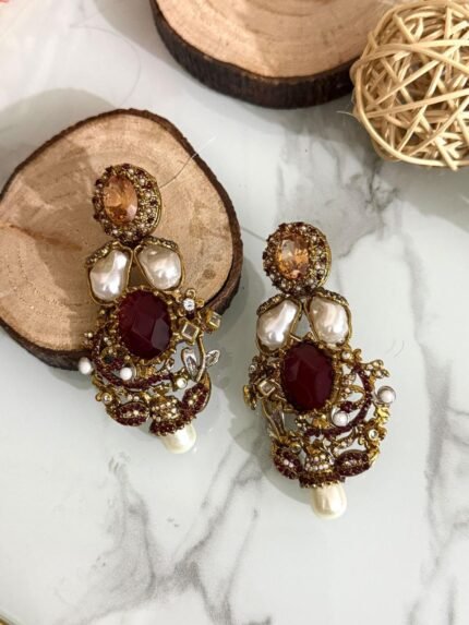 A rich collection of ornate drop earrings featuring oversized gemstones, baroque-style pearls, and detailed gold filigree with colored stone accents — arranged on a marble surface with natural wood and wicker decor.