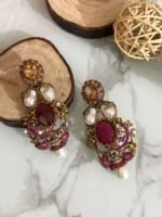 A rich collection of ornate drop earrings featuring oversized gemstones, baroque-style pearls, and detailed gold filigree with colored stone accents — arranged on a marble surface with natural wood and wicker decor.