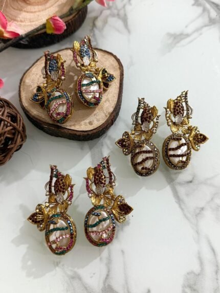 A whimsical collection of handcrafted earrings featuring oval faux pearls with colorful mosaic-like gem patterns, gold-toned floral accents, and intricate detailing, arranged on a marble surface with wood and floral props.