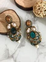 A rich collection of ornate drop earrings featuring oversized gemstones, baroque-style pearls, and detailed gold filigree with colored stone accents — arranged on a marble surface with natural wood and wicker decor.