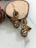 A whimsical collection of handcrafted earrings featuring oval faux pearls with colorful mosaic-like gem patterns, gold-toned floral accents, and intricate detailing, arranged on a marble surface with wood and floral props.