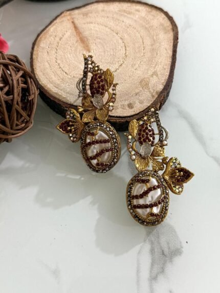 A whimsical collection of handcrafted earrings featuring oval faux pearls with colorful mosaic-like gem patterns, gold-toned floral accents, and intricate detailing, arranged on a marble surface with wood and floral props.