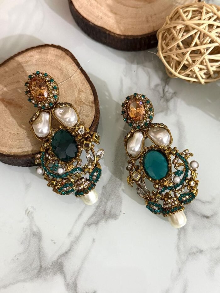 A rich collection of ornate drop earrings featuring oversized gemstones, baroque-style pearls, and detailed gold filigree with colored stone accents — arranged on a marble surface with natural wood and wicker decor.
