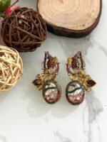 A whimsical collection of handcrafted earrings featuring oval faux pearls with colorful mosaic-like gem patterns, gold-toned floral accents, and intricate detailing, arranged on a marble surface with wood and floral props.