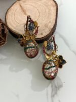 A whimsical collection of handcrafted earrings featuring oval faux pearls with colorful mosaic-like gem patterns, gold-toned floral accents, and intricate detailing, arranged on a marble surface with wood and floral props.